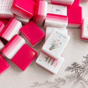 May include: A collection of mahjong tiles featuring a vibrant hot pink and white color scheme. Several tiles display intricate illustrations, including a flower with the text FLOWER 8, a red dragon symbol with the text RED, and a green pagoda.