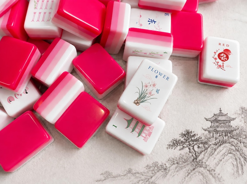 May include: A collection of mahjong tiles featuring a vibrant hot pink and white color scheme. Several tiles display intricate illustrations, including a flower with the text FLOWER 8, a red dragon symbol with the text RED, and a green pagoda.