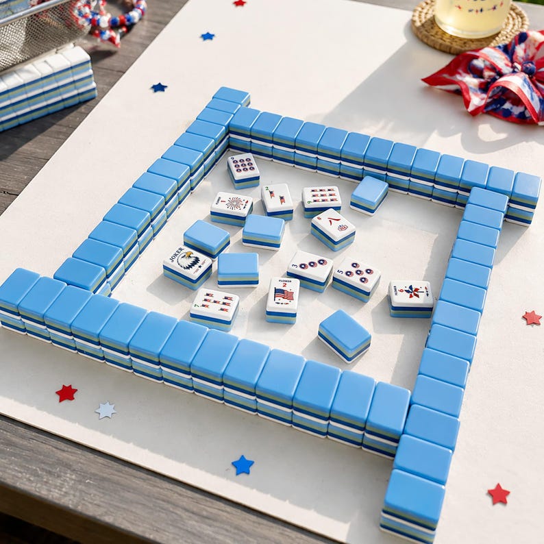 May include: A Mahjong set featuring light blue tiles with white and green accents. The tiles are arranged in a square perimeter with scattered tiles in the center, displaying various symbols including an American flag, a bald eagle, and numbers.