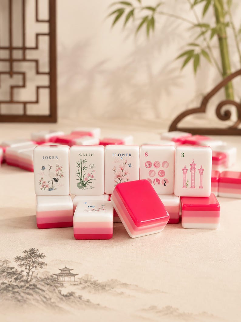 Op de afbeelding: Een set witte mahjongtegels met roze gradi&euml;ntvoeten. De tegels tonen diverse illustraties waaronder een kraanvogel, bamboe, bloemen en pagodes, met tekstlabels zoals JOKER, GREEN, FLOWER 5, 8 en 3 gedrukt in zwarte of roze inkt.