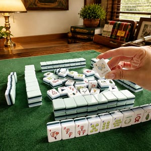 May include: A Mahjong set featuring rectangular tiles with light green backs and white faces. The tiles display various colorful illustrations, including numbers, floral motifs, and the word FLOWER. Some tiles are arranged in rows on a green surface.