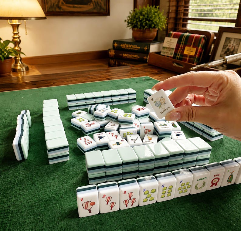 May include: A Mahjong set featuring rectangular tiles with light green backs and white faces. The tiles display various colorful illustrations, including numbers, floral motifs, and the word FLOWER. Some tiles are arranged in rows on a green surface.