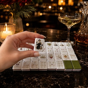 May include: A white rectangular mahjong tile featuring a graphic illustration of a woman in a black dress holding a cocktail glass with the text JOKER printed below. The tile is held above a set of matching tiles with various cocktail illustrations.