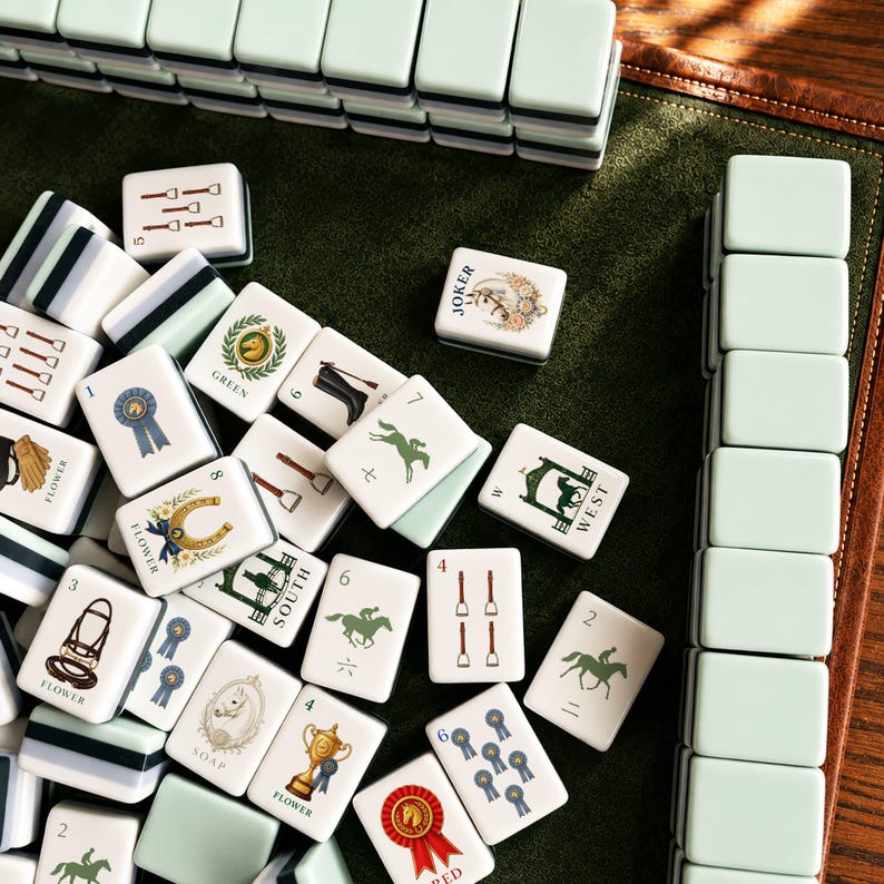 May include: A set of mahjong tiles featuring light green backs and white faces with various equestrian-themed illustrations, including horses, trophies, ribbons, and text labels like Joker, Green, West, South, Flower, Soap, and Red, arranged on a green surface.