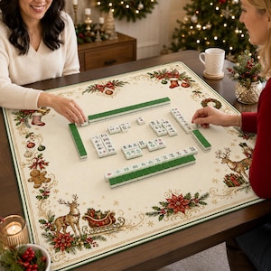 May include: A square mahjong mat featuring a cream-colored surface with a festive Christmas border print including reindeer, poinsettias, gingerbread men, and stockings. The mat is set with white and green mahjong tiles in a game arrangement.