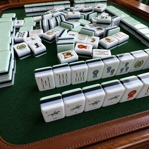 May include: A set of white mahjong tiles with light green, black, and white striped backs, featuring various equestrian-themed illustrations, numbers, and text such as Green, Flower, and Red, arranged on a dark green felt surface with a brown border.