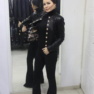 May include: A black leather jacket with gold buttons is worn over a black turtleneck and black flared pants. The jacket's interior features a patterned lining. The outfit is displayed in a clothing store.