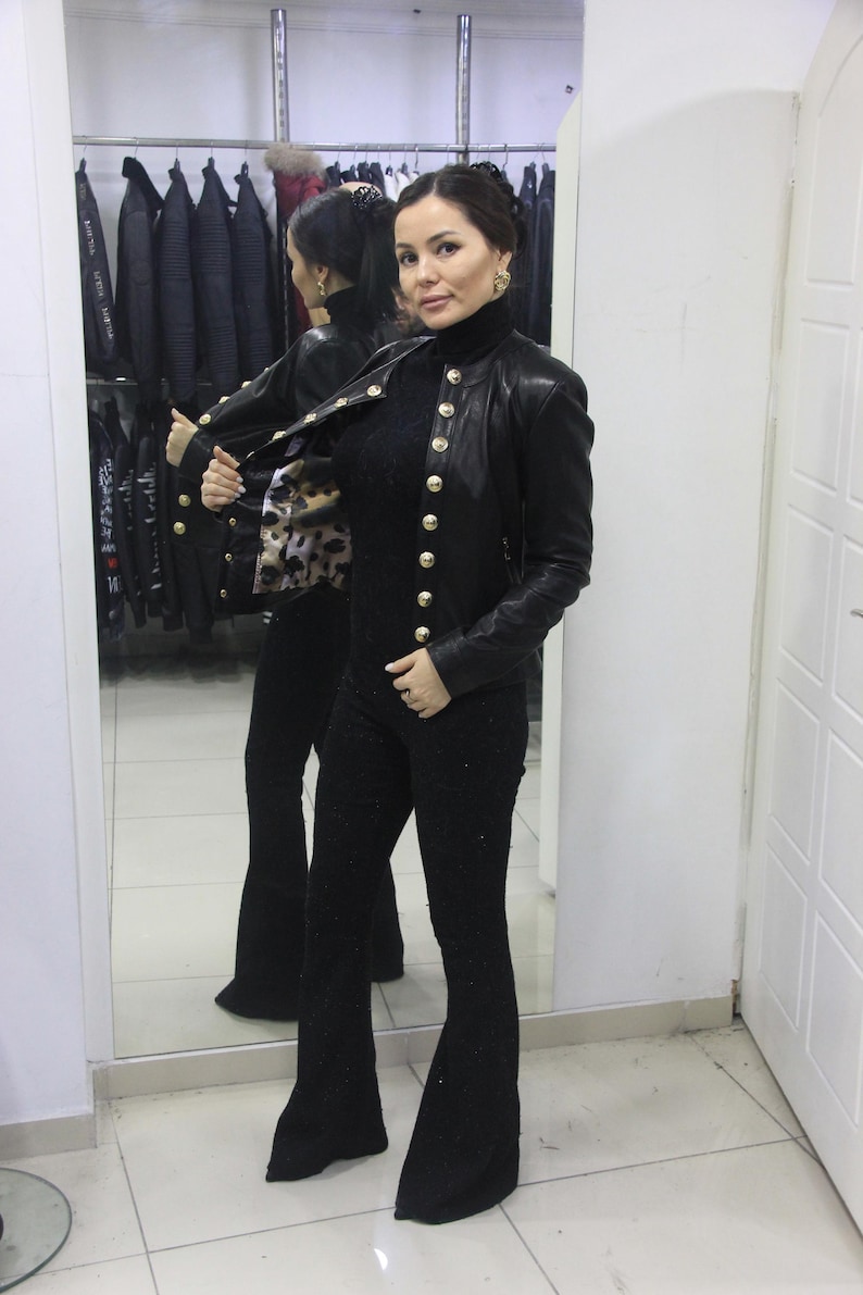 May include: A black leather jacket with gold buttons is worn over a black turtleneck and black flared pants. The jacket's interior features a patterned lining. The outfit is displayed in a clothing store.