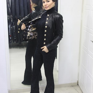 May include: A black leather jacket with gold buttons is worn over a black outfit. The jacket has a classic design with a high collar. The outfit includes a black top and flared pants. A leopard print scarf is visible.