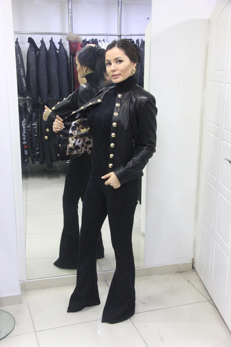 May include: A black leather jacket with gold buttons is worn over a black outfit. The jacket has a classic design with a high collar. The outfit includes a black top and flared pants. A leopard print scarf is visible.