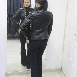 May include: A woman in a black leather jacket with gold buttons and black pants is reflected in a mirror. The jacket has a high collar and long sleeves. The pants are flared. A clothing rack is visible in the background.