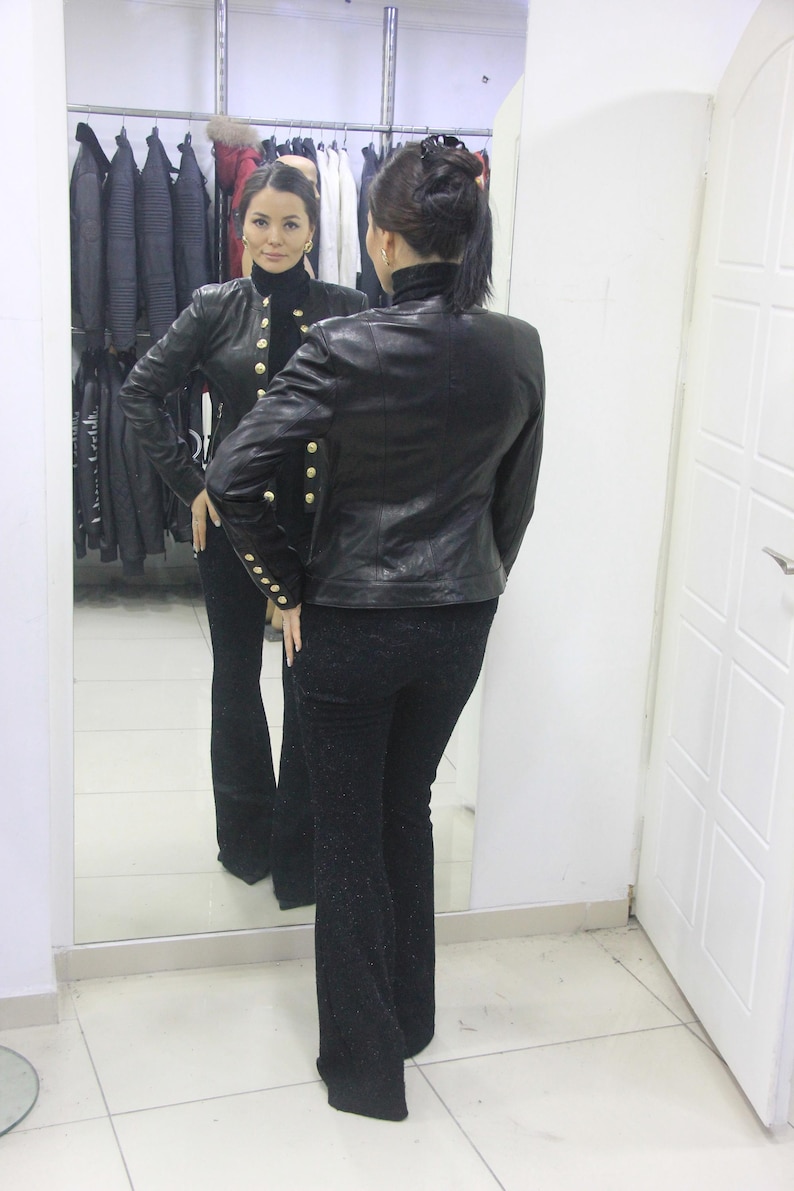 May include: A woman in a black leather jacket with gold buttons and black pants is reflected in a mirror. The jacket has a high collar and long sleeves. The pants are flared. A clothing rack is visible in the background.