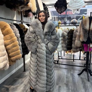 May include: A woman wearing a long, gray fur coat. The coat has a large collar and a full, floor-length design. Other fur coats in various colors hang on racks in the background, along with fur hats.