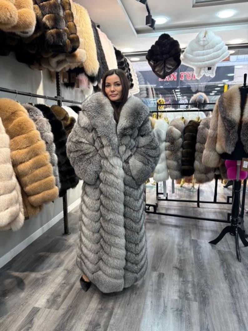 May include: A woman wearing a long, gray fur coat. The coat has a large collar and a full, floor-length design. Other fur coats in various colors hang on racks in the background, along with fur hats.