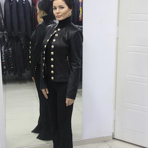 May include: A black leather jacket with gold buttons and a high collar is paired with black flared pants. The jacket has multiple gold buttons down the front. The outfit is displayed in a clothing store setting.