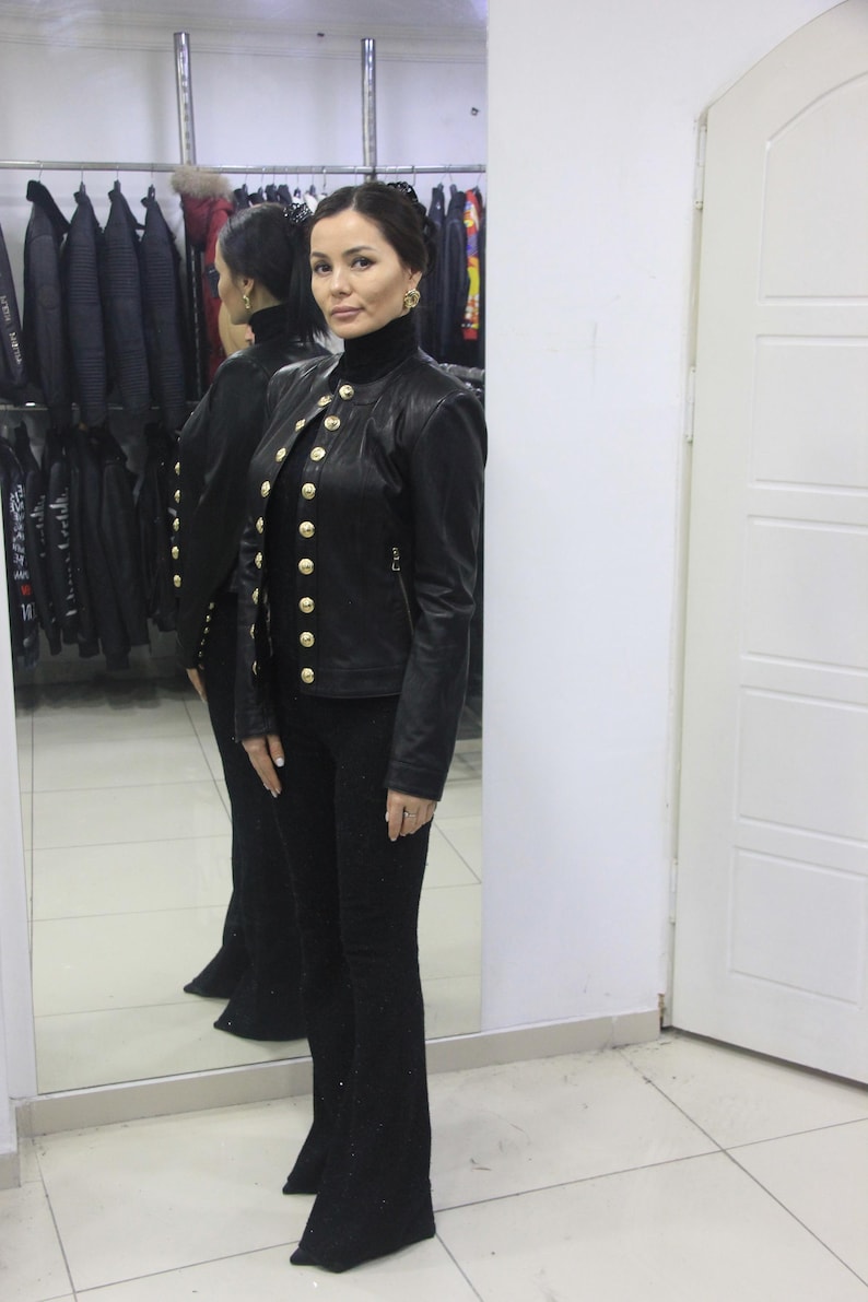 May include: A black leather jacket with gold buttons and a high collar is paired with black flared pants. The jacket has multiple gold buttons down the front. The outfit is displayed in a clothing store setting.