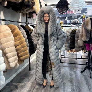 May include: A woman wearing a long, silver-gray fur coat with a large hood. The coat is floor-length and has a textured appearance. Other fur coats in various colors hang on racks in the background, showcasing a collection of outerwear.