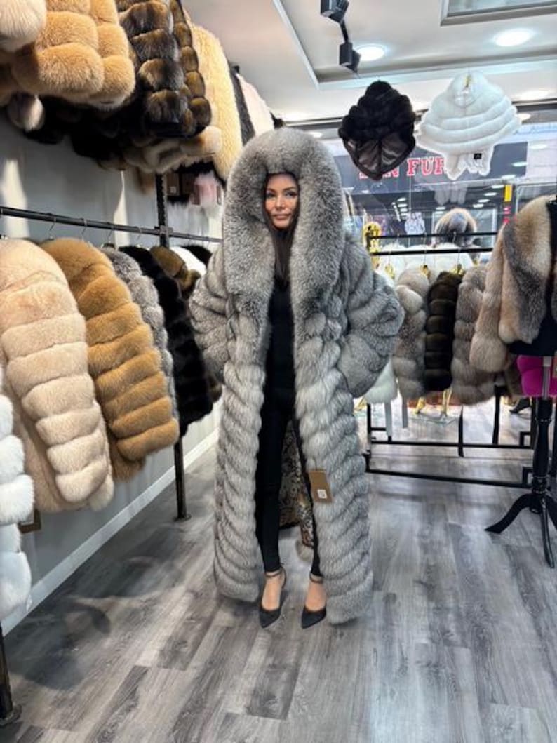 May include: A woman wearing a long, silver-gray fur coat with a large hood. The coat is floor-length and has a textured appearance. Other fur coats in various colors hang on racks in the background, showcasing a collection of outerwear.