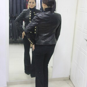 May include: A black leather jacket with gold buttons and matching black pants are reflected in a mirror. The jacket has a high collar and long sleeves. The pants are flared at the bottom.
