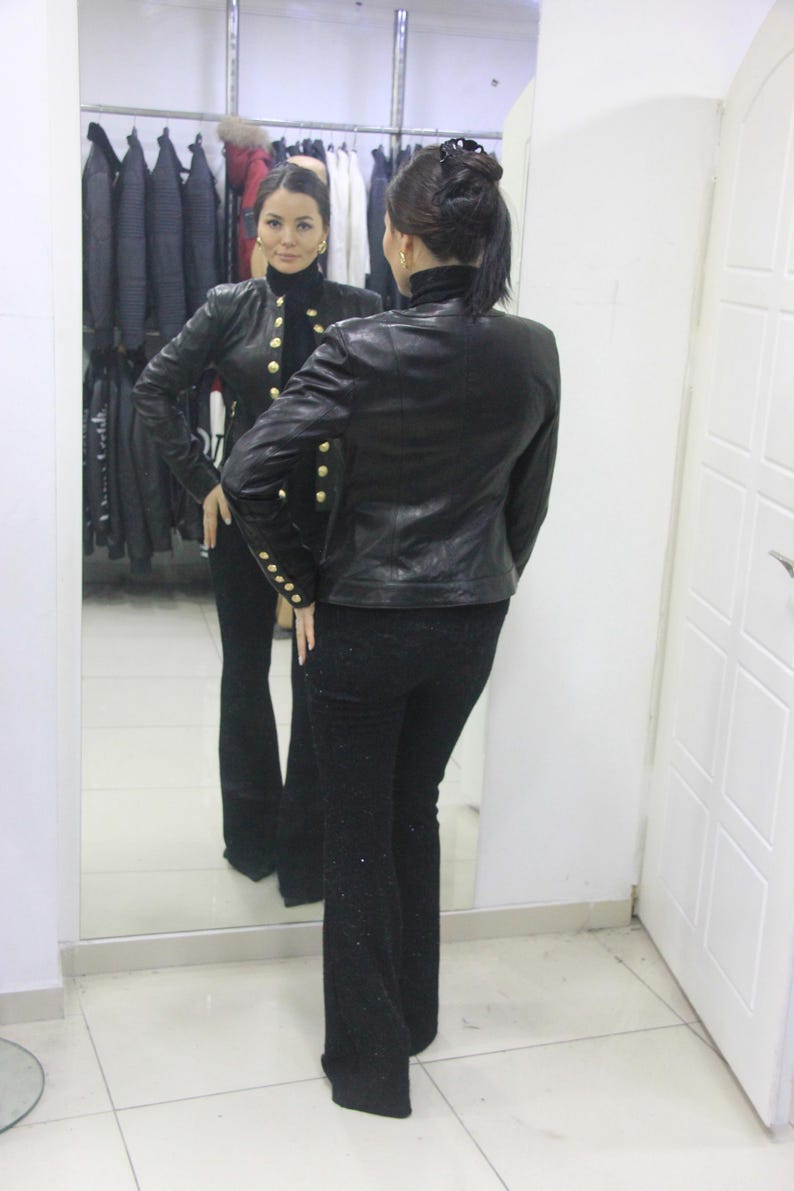 May include: A black leather jacket with gold buttons and matching black pants are reflected in a mirror. The jacket has a high collar and long sleeves. The pants are flared at the bottom.