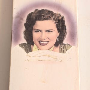 May include: A Patsy Cline Collection box set featuring a portrait of the singer. The box is primarily white with the title "Patsy Cline" in large, stylized letters at the top. The image of Cline is in a purple oval.