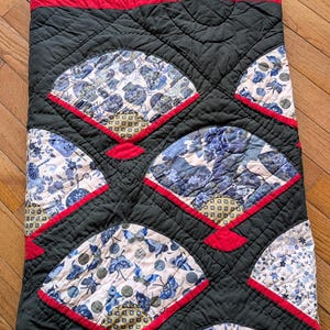 May include: A handmade quilt featuring a repeating fan pattern. The fans are filled with blue and white floral fabric, outlined in red, and set against a dark gray background. The top border has a blue floral pattern.