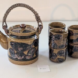May include: A ceramic tea set featuring a teapot with a woven handle and lid, and four matching cups. The set has a brown and black floral design. The teapot has a brown spout and lid. The cups are stacked in pairs.