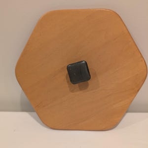 May include: A light brown, hexagonal wooden tray with a small, dark grey square handle in the centre. The tray has a smooth surface and a natural wood grain pattern. The tray is on a white surface.