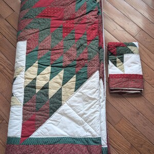 May include: A folded quilt and a smaller folded quilt, both featuring a patchwork design. The quilts have a color scheme of red, green, white, and tan. The larger quilt is folded and has a patterned border.