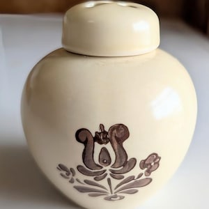 May include: Cream-colored ceramic jar with a matching lid. The jar features a brown floral design on the front. The lid has small holes. The jar has a rounded shape.