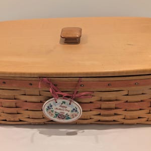 May include: A rectangular woven basket with a wooden lid and handle. The basket is light brown with a pink accent band. A decorative tag with floral details and the text "Happy Mother's Day" is attached.