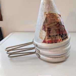May include: A stack of three silver-colored rotary food mills with handles, topped by a perforated cone-shaped strainer. The strainer has a partially visible label with the words "Rotary Food Mill" and "Comet".