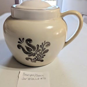 May include: Cream-colored ceramic bean pot or cookie jar with a lid and handle. The jar features a dark gray floral design on the front. The text on the paper reads "Bean pot/Cookie Jar With Lid - #70" and "#81".