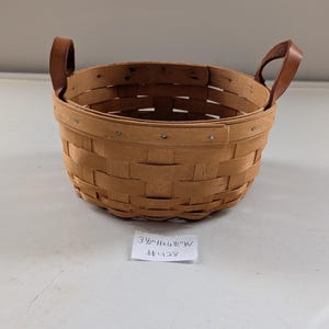 May include: A round, woven basket with a light brown hue. It features two brown leather handles and is labeled with dimensions of 3.5"H x 6"W. The basket is made of woven wood strips and is held together with small metal fasteners.
