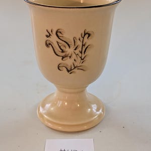 May include: A beige ceramic goblet with a black floral design and a black rim. The goblet has a pedestal base and a flared top. The text "#475" is on a white label below the goblet.