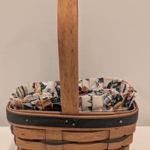 May include: A woven basket with a wooden handle and a floral fabric lining. The basket is made of light brown wood with dark gray accents. The fabric lining features a pattern of flowers in various colors.