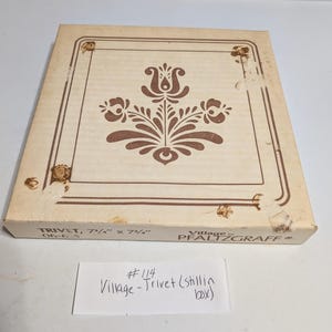 May include: A square trivet in its original box, featuring a brown floral design on a beige background. The box is labeled "TRIVET, 7 3/4" x 7 3/4"" and "Village by PFALTZGRAFF". A handwritten note reads "#114 Village-Trivet (still in box)".