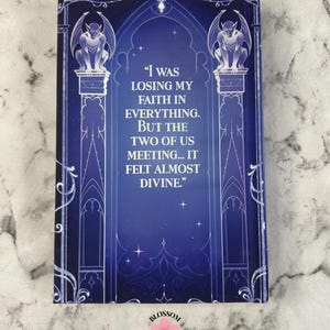 Puede incluir: Una portada de libro con un fondo azul oscuro y detalles en blanco. La portada presenta una cita: "Estaba perdiendo la fe en todo. Pero nosotros dos reuni&eacute;ndonos... se sinti&oacute; casi divino." Dos g&aacute;rgolas se sientan sobre columnas. El logotipo de Blossom Binds est&aacute; en la parte inferior.