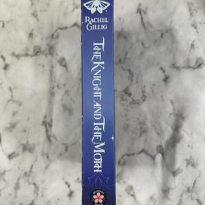 Puede incluir: Lomo de libro azul con el t&iacute;tulo "The Knight and The Moth" en escritura blanca. El nombre del autor, Rachel Gillig, est&aacute; encima del t&iacute;tulo. Un dise&ntilde;o de polilla y floral adorna la parte superior e inferior del lomo.