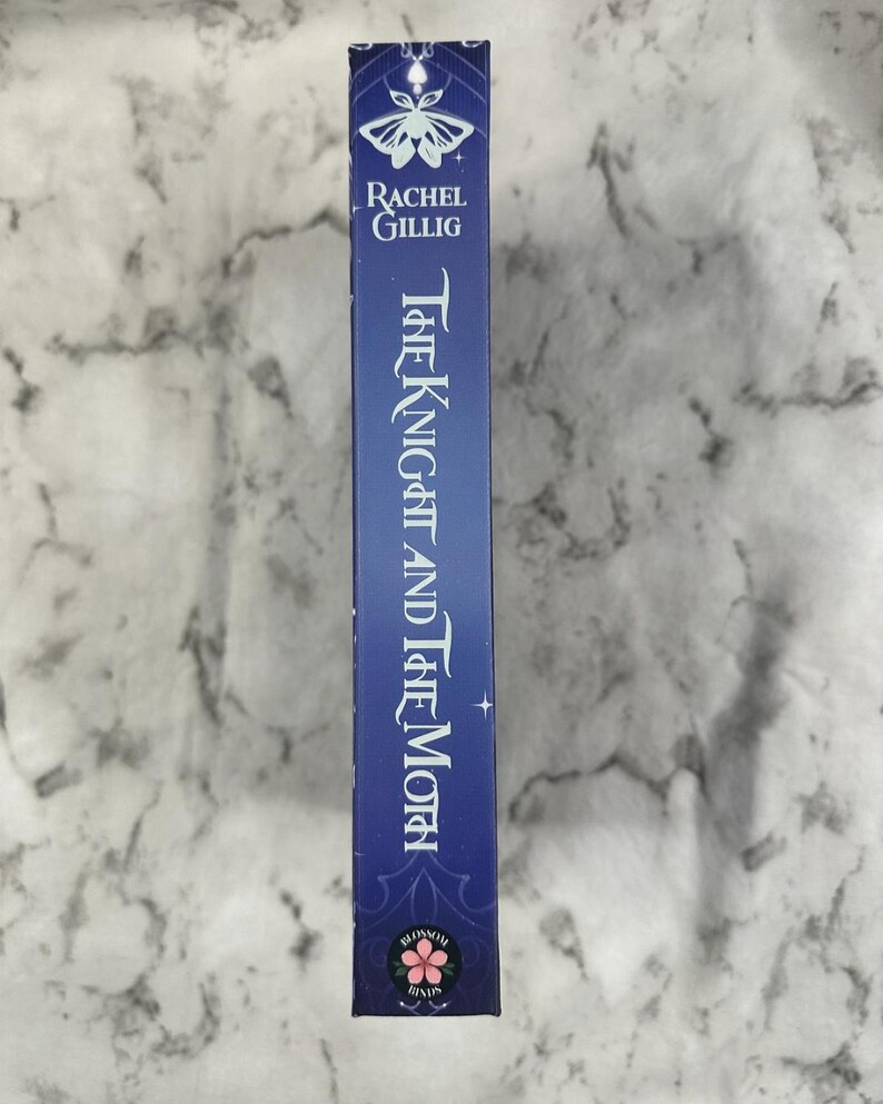 Puede incluir: Lomo de libro azul con el t&iacute;tulo "The Knight and The Moth" en escritura blanca. El nombre del autor, Rachel Gillig, est&aacute; encima del t&iacute;tulo. Un dise&ntilde;o de polilla y floral adorna la parte superior e inferior del lomo.