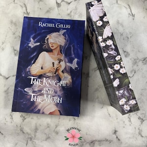 Puede incluir: Un libro titulado "The Knight and The Moth" de Rachel Gillig. La portada presenta a una mujer con los ojos vendados, sosteniendo un martillo, con mariposas y un fondo azul. El lomo del libro est&aacute; decorado con mariposas blancas y flores.