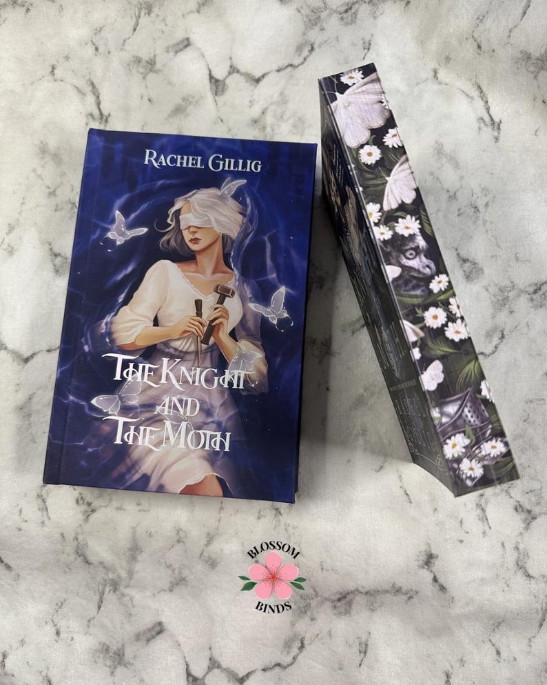 Puede incluir: Un libro titulado "The Knight and The Moth" de Rachel Gillig. La portada presenta a una mujer con los ojos vendados, sosteniendo un martillo, con mariposas y un fondo azul. El lomo del libro est&aacute; decorado con mariposas blancas y flores.