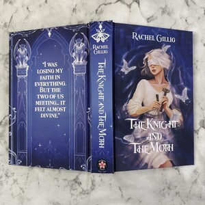 Puede incluir: Libro de tapa dura titulado "The Knight and The Moth" de Rachel Gillig. La portada presenta a una mujer con los ojos vendados, sosteniendo herramientas y mariposas. El lomo y la contraportada son de color azul oscuro con elementos decorativos y texto.