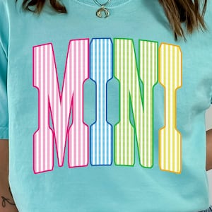 Puede incluir: Una camiseta azul claro con la palabra "MINI" en letras grandes y coloridas. Las letras están rayadas verticalmente en rosa, azul, verde y amarillo, con contornos blancos. La camiseta tiene mangas cortas y cuello redondo.