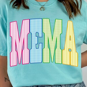 May include: A light blue t-shirt with the word "MEMA" in large, colorful, striped letters. The letters are pink, blue, green, and yellow. The shirt has short sleeves and a crew neck.