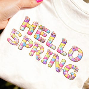 Könnte beinhalten: Ein cremefarbenes T-Shirt mit dem Schriftzug "HELLO SPRING" in bunten Buchstaben im Patchwork-Stil. Jeder Buchstabe ist mit Schmetterlingen und floralen Akzenten verziert, was ein fröhliches Frühlingsdesign ergibt.