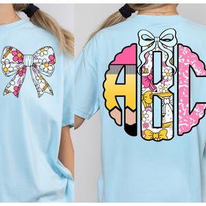 May include: A light blue t-shirt featuring a graphic of a bow with a daisy and pencil pattern on the front, and a large circular monogram with the letters ABC, decorated with pencils, daisies, and a bow on the back of the garment.
