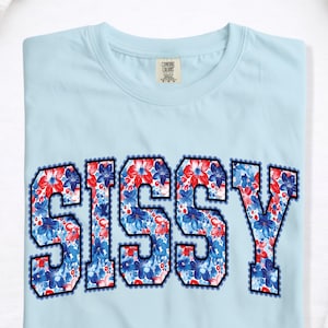May include: Light blue t-shirt with the word "SISSY" in large, bold letters. The letters are filled with a floral pattern in red and blue, outlined in black with a scalloped edge. The shirt has a crew neck.