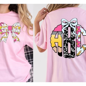 Op de afbeelding: Een lichtroze t-shirt met een strik-afbeelding op de voorkant en een groot cirkelvormig monogram met de letters ABC op de achterkant. De strik en het monogram zijn versierd met patronen van gele potloden, roze bloemen en zwart-witte texturen.