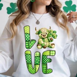 May include: White sweatshirt with the word "LOVE" in green patchwork letters. A patchwork Highland cow holding a shamrock is between the letters. The sweatshirt is decorated with green polka dots, stripes, and plaid patterns, perfect for St. Patrick's Day.
